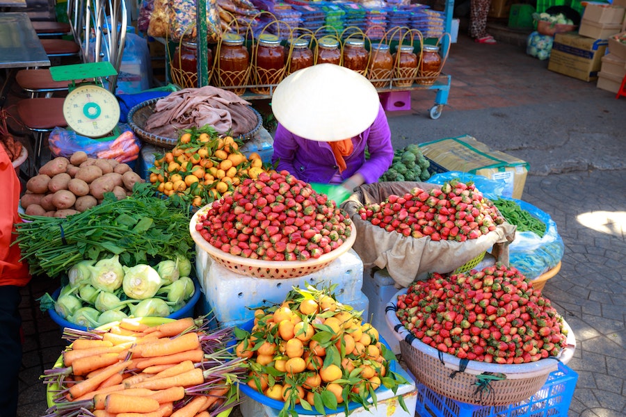 Asian woman in cone hat in front of vegetable piles at market